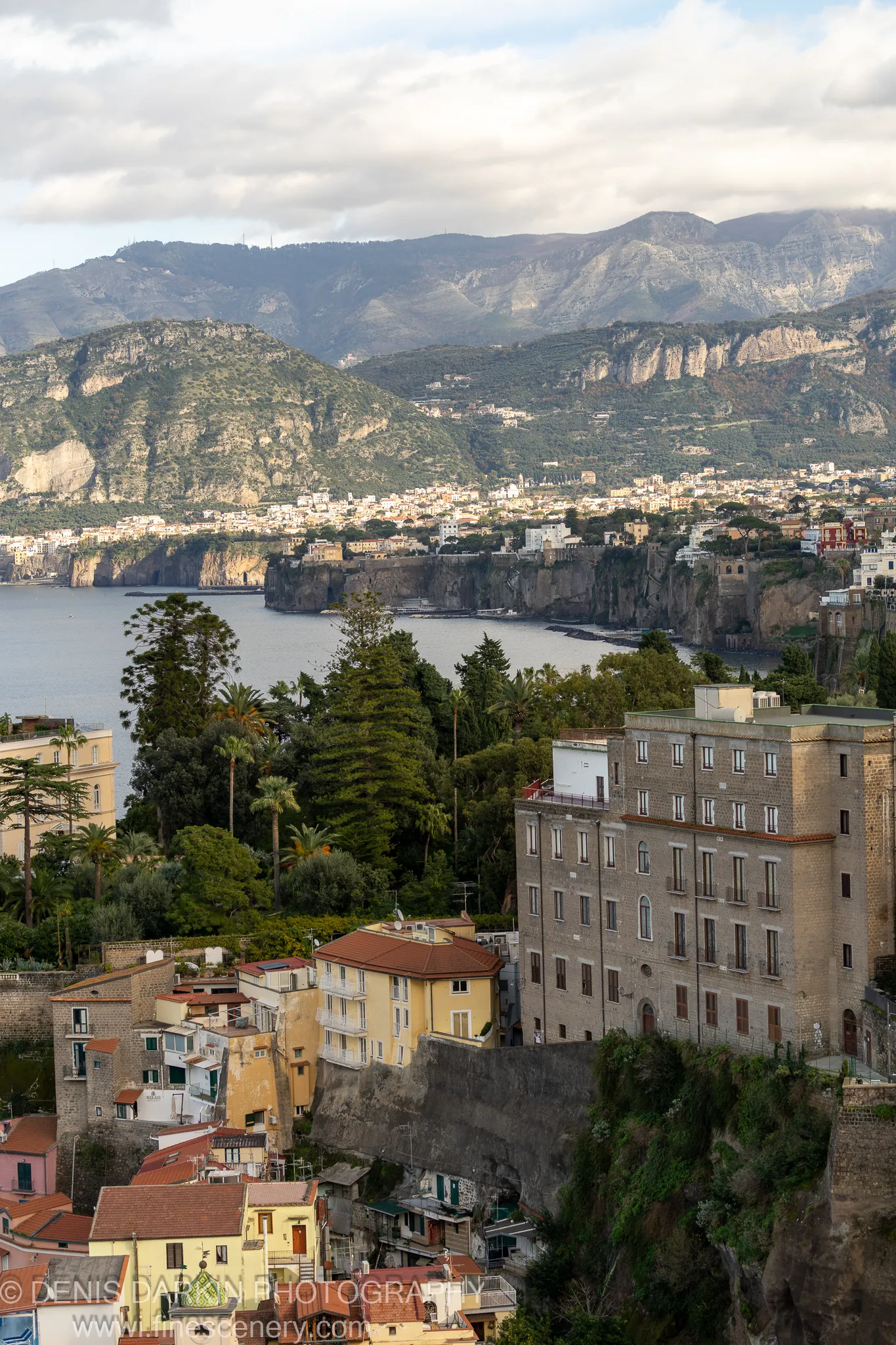 Terracotta-roofed buildings on a steep Amalfi Coast slope, stone walls in soft directional light, a calm bay and distant mountains beyond. December 2024.