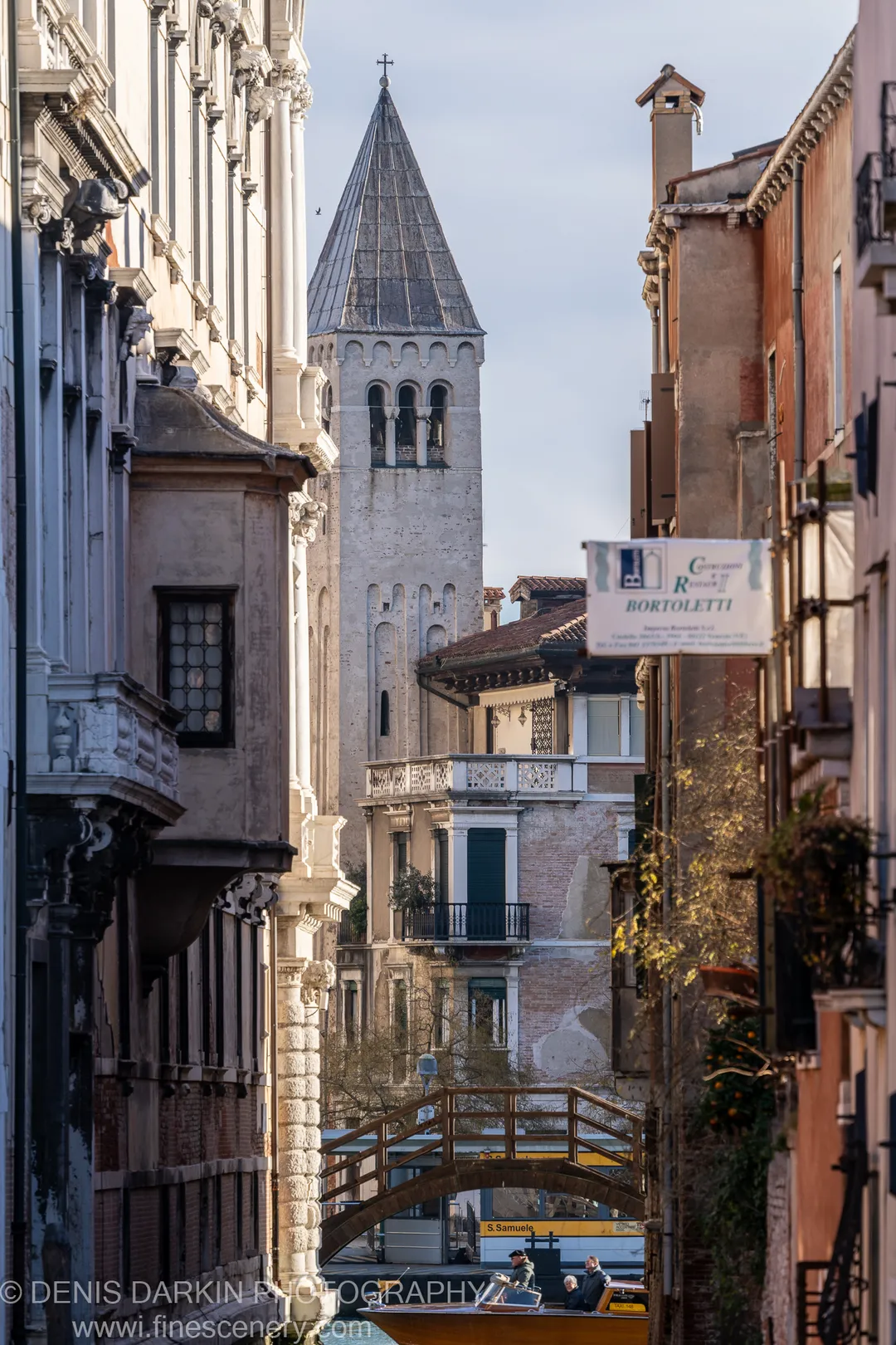 Textured stone facades flank a narrow Venetian canal, with a wooden footbridge below and a yellow water taxi heading toward a distant church tower.