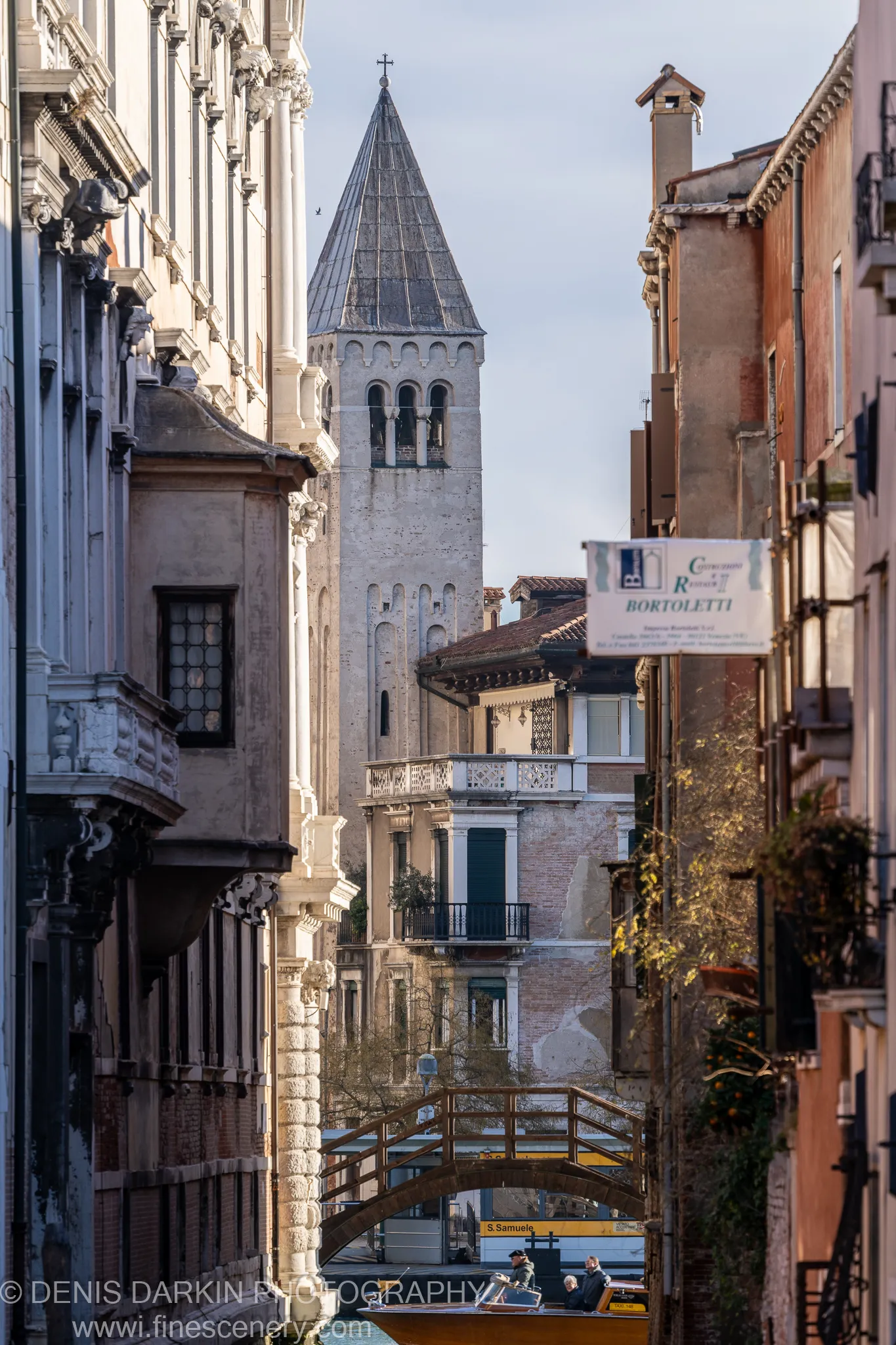 Textured stone facades flank a narrow Venetian canal, with a wooden footbridge below and a yellow water taxi heading toward a distant church tower.