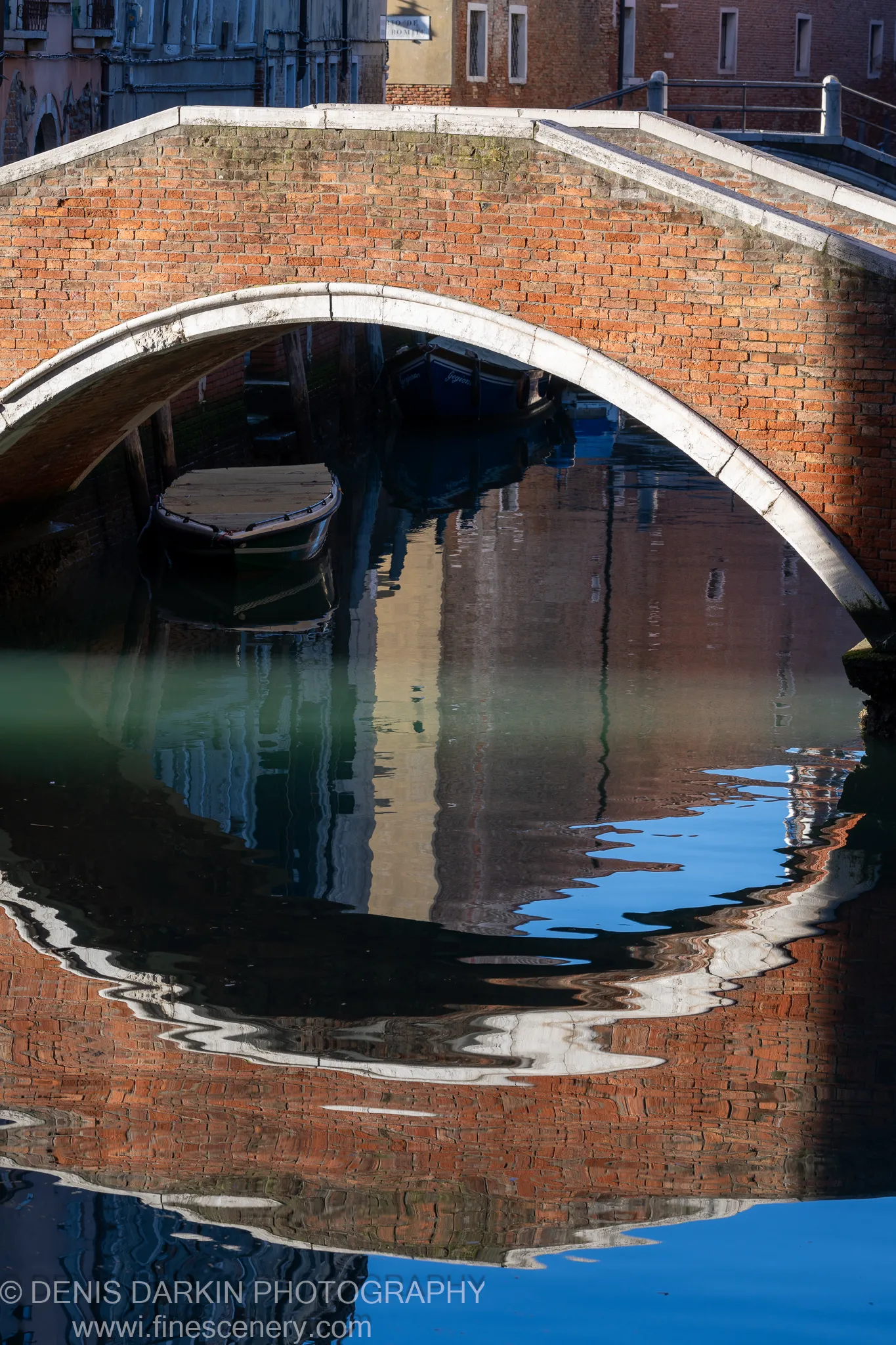 A weathered red brick arch spans a calm Venetian canal above a moored boat, with warm building reflections distorted in the water below.