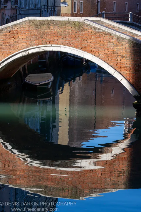 A weathered red brick arch spans a calm Venetian canal above a moored boat, with warm building reflections distorted in the water below.