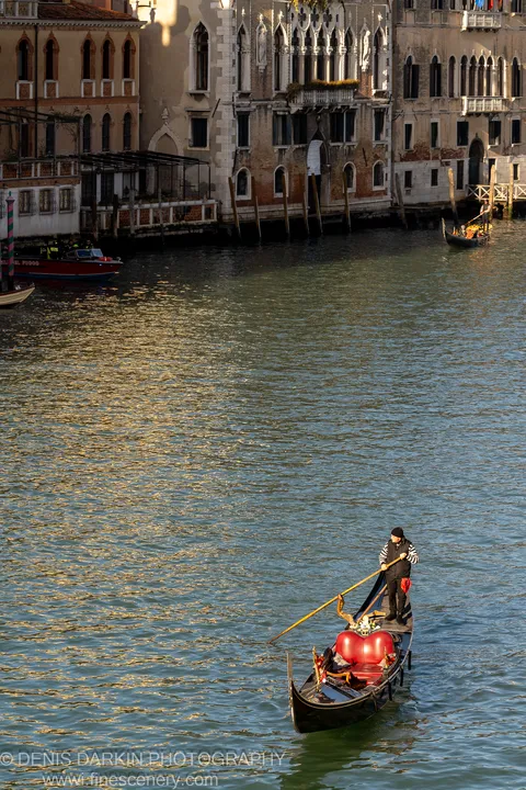 A gondola with a red interior sits on rippling canal water in Venice, with Gothic-arched building facades lining the far bank in soft morning light.