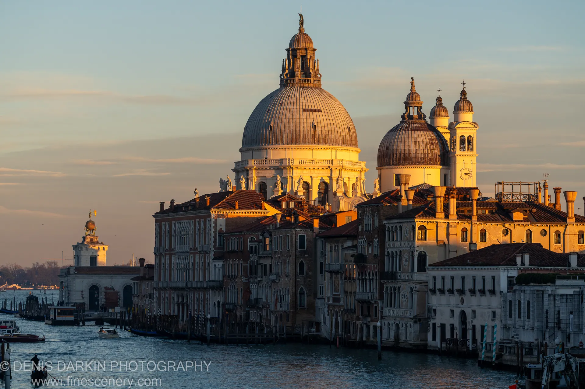 Ornate terracotta rooftops and stone facades along a Venice canal waterfront catch warm afternoon light beneath a pale winter sky with scattered clouds.