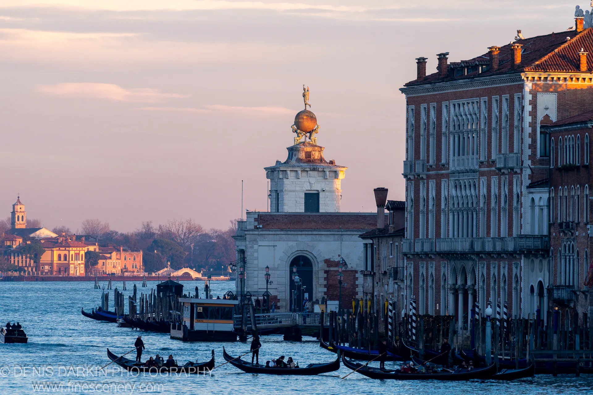 Gondoliers navigate a Venice canal at sunset, amber light on brick facades contrasting with cool blue water below a tower with a golden sphere.