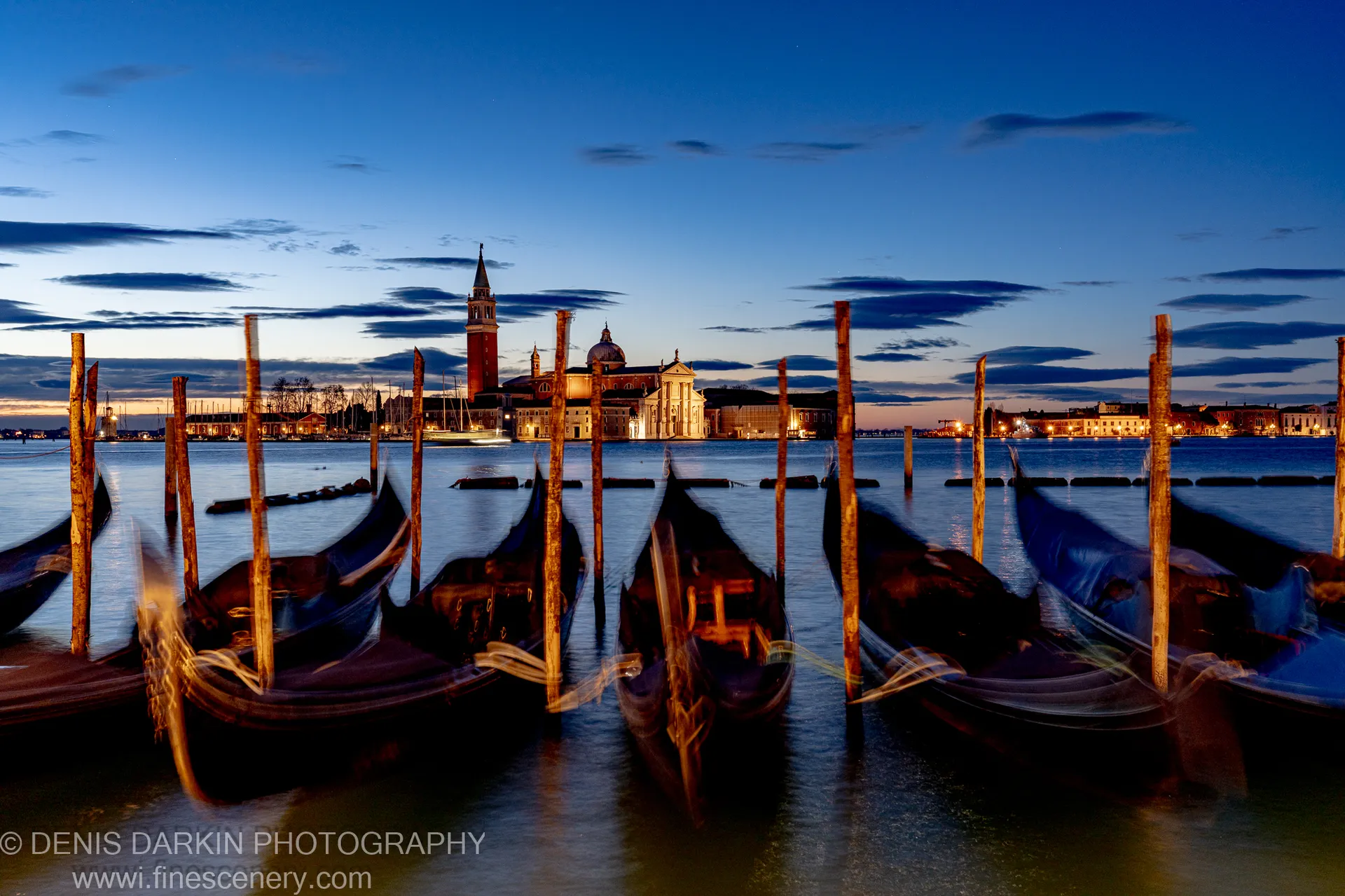 Vertical wooden pilings cross a calm Venice harbor at dawn, leading toward a softly lit church complex and bell tower on the horizon.