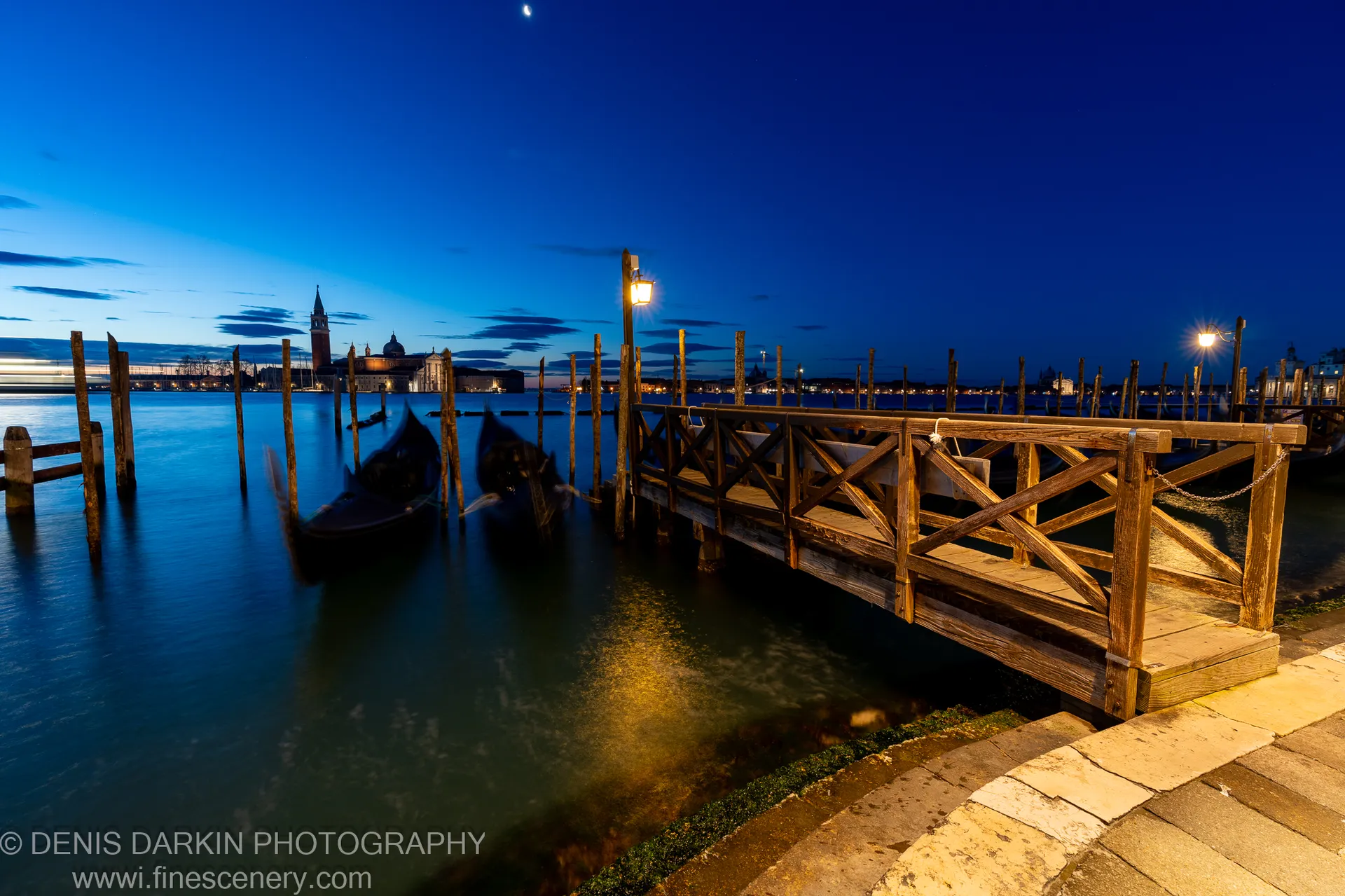 A wooden pier and weathered pilings at a Venice waterfront at dawn, harbor lights reflected in still water, a crescent moon above.