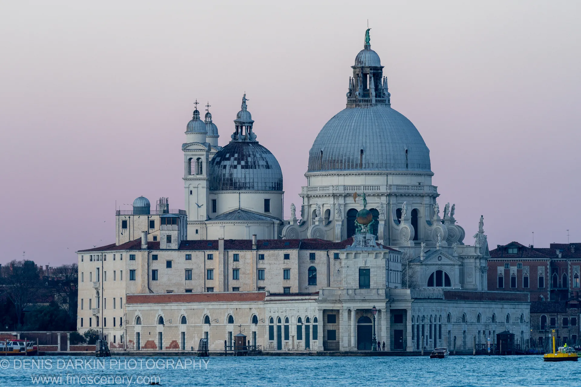 A grand domed basilica rises above a Venice waterfront promenade under a soft violet dawn sky, cool blue lagoon water in the foreground.