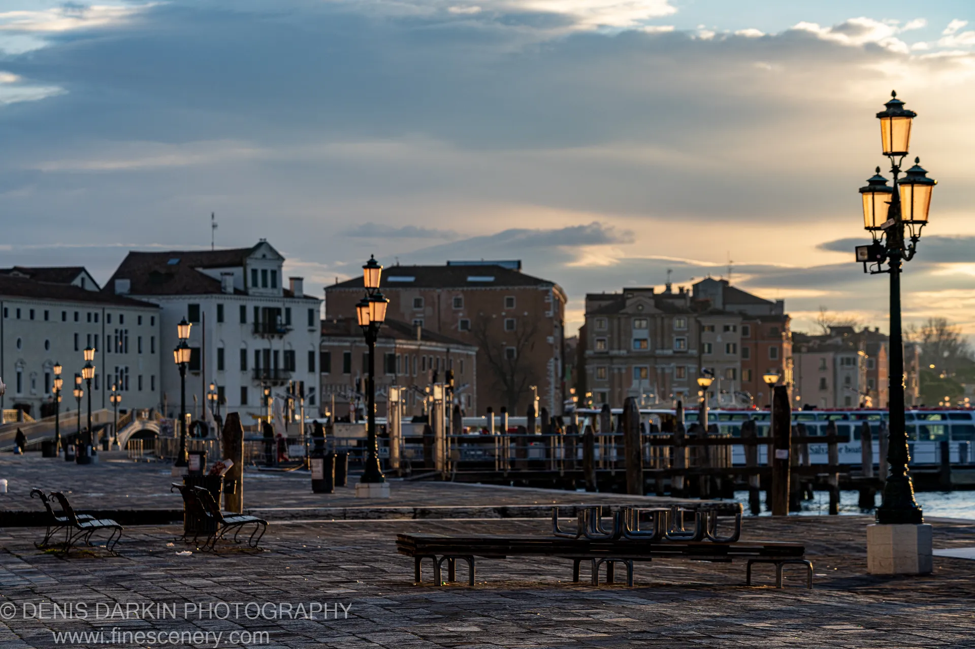Classic glass streetlamps on a cobblestone Venice waterfront plaza at dawn, a marina and layered facades compressed in the distance under grey cloud.