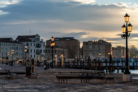 Classic glass streetlamps on a cobblestone Venice waterfront plaza at dawn, a marina and layered facades compressed in the distance under grey cloud.