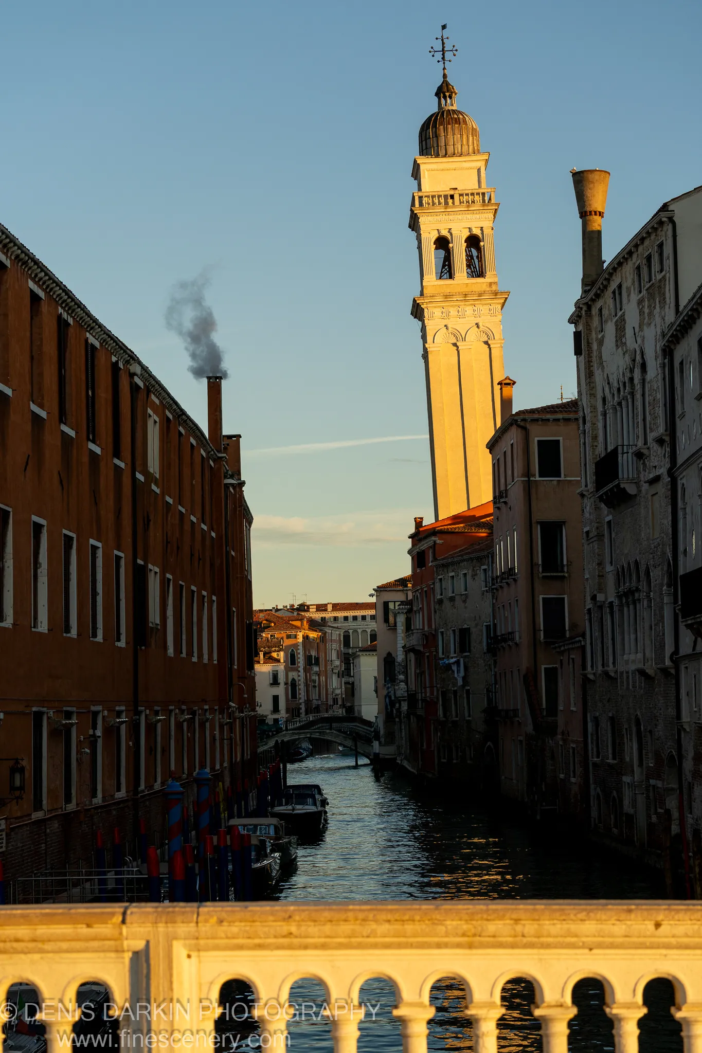 Dawn light catches the top of a Venetian campanile above a stone balustrade, while chimney smoke drifts across the blue sky.