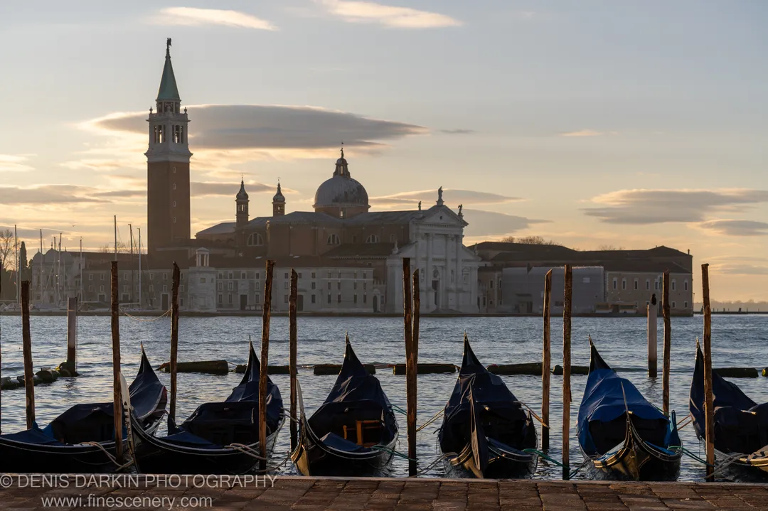 Covered gondolas moored along weathered wooden poles in the foreground, with a church campanile and dome lit by dawn light beyond.