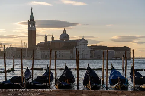 Covered gondolas moored along weathered wooden poles in the foreground, with a church campanile and dome lit by dawn light beyond.