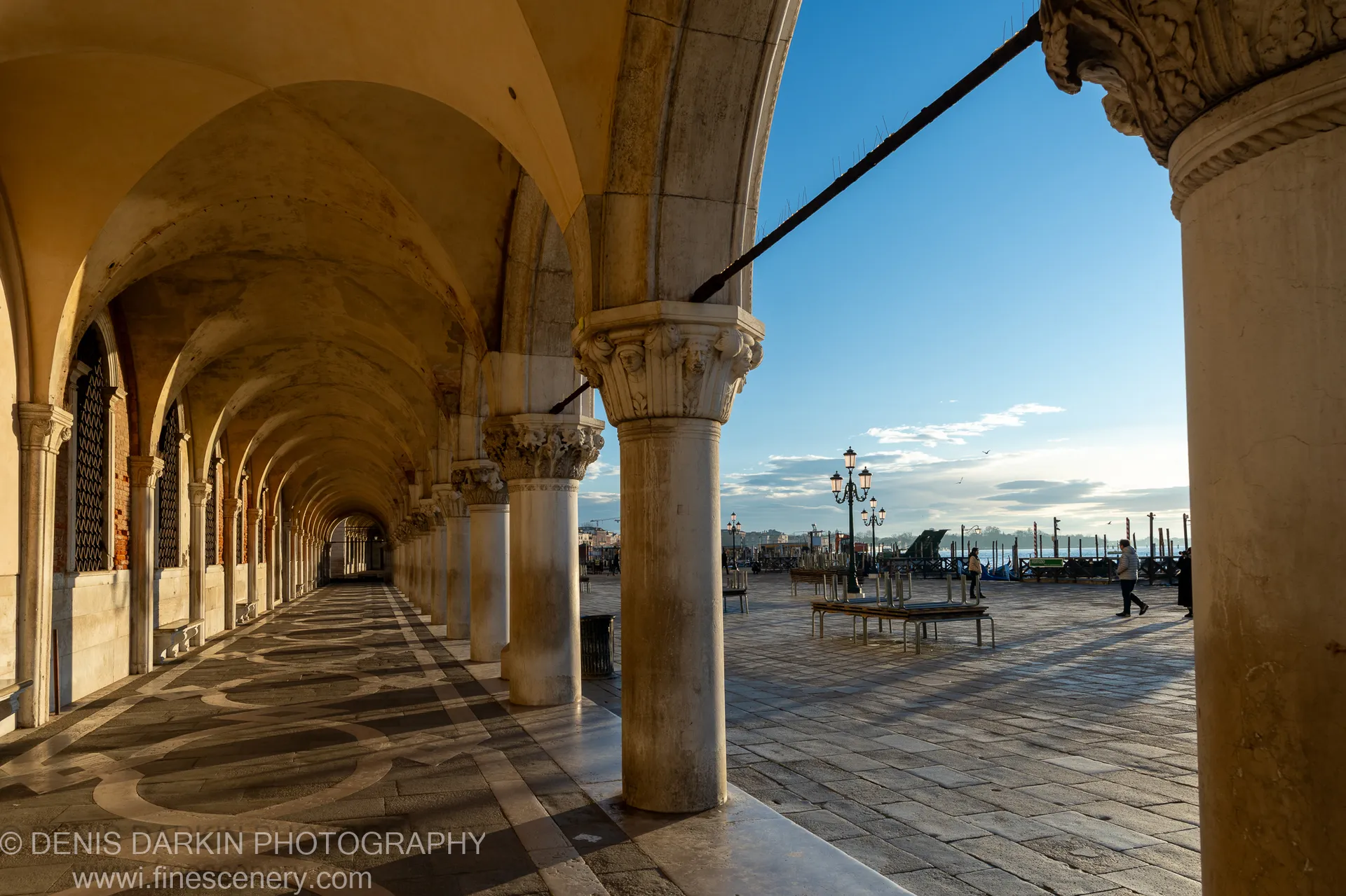 Long perspective through a stone arcade, sunlight casting geometric shadows across patterned pavement toward a blue sky and waterfront.