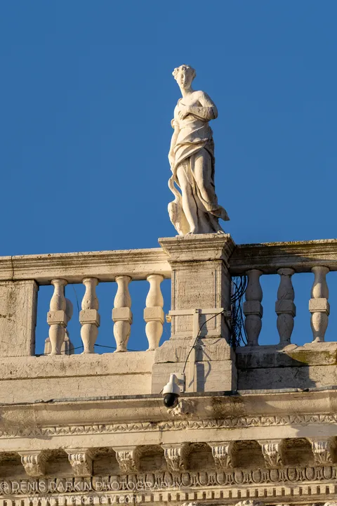 Weathered marble statue on a balustrade against deep blue sky, a security camera mounted on the wall below.
