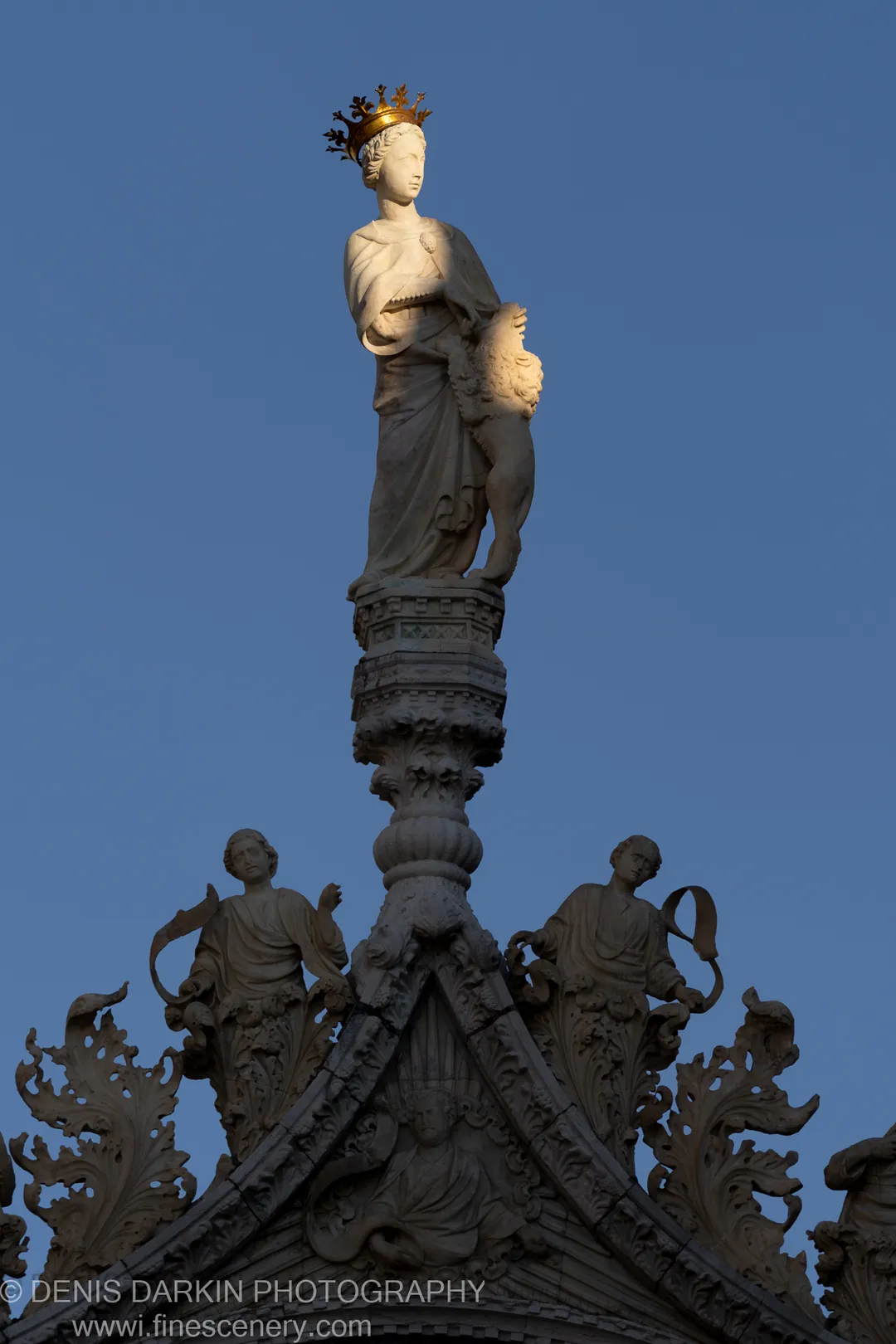 Upward view of a crowned stone statue at the apex of a Venetian facade, with a carved lion beside it against blue sky.