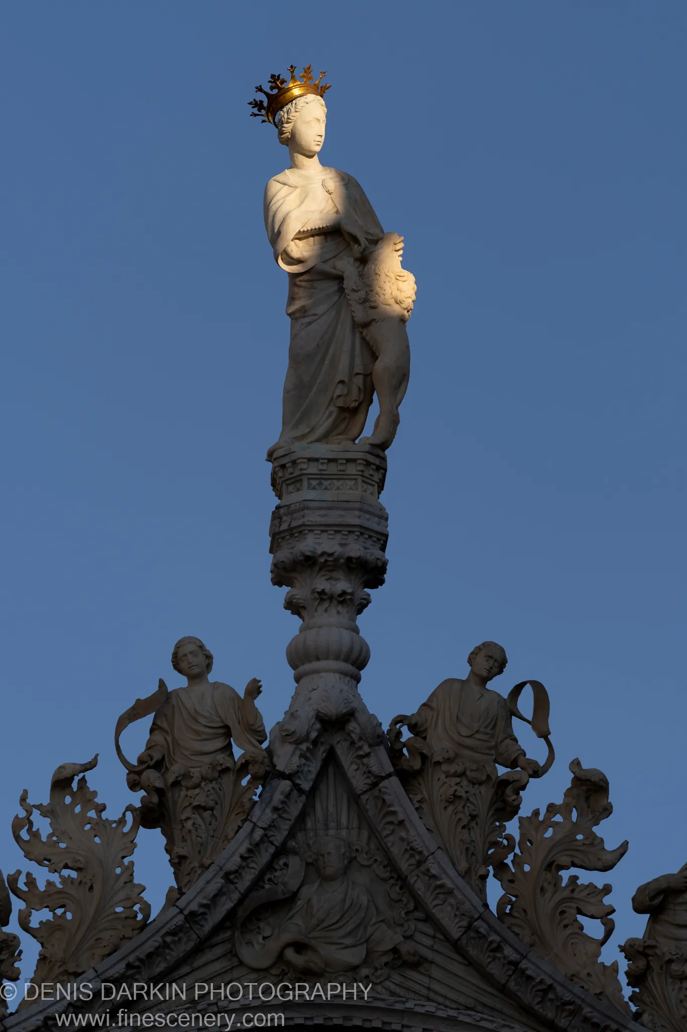 Upward view of a crowned stone statue at the apex of a Venetian facade, with a carved lion beside it against blue sky.