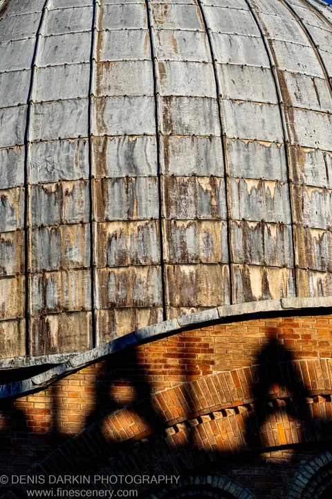 Close detail of a Venetian dome where grey metal cladding shows vertical rust streaks above courses of warm terracotta brick in sharp morning light.