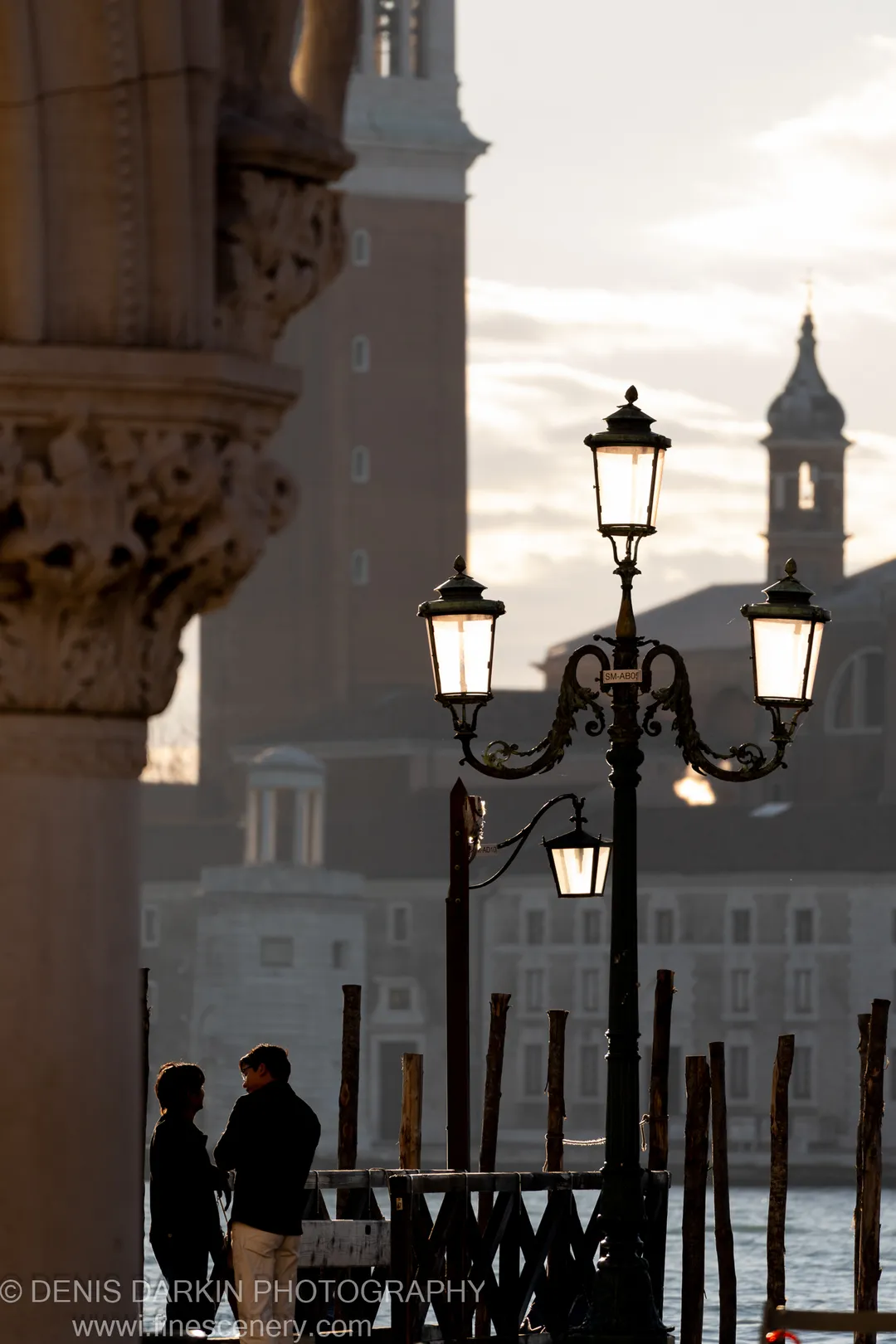 A three-lantern iron post on the Venice waterfront, a stone column to the left and church towers in soft silhouette beyond.