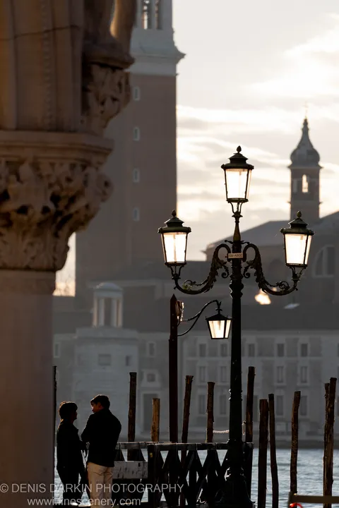 A three-lantern iron post on the Venice waterfront, a stone column to the left and church towers in soft silhouette beyond.