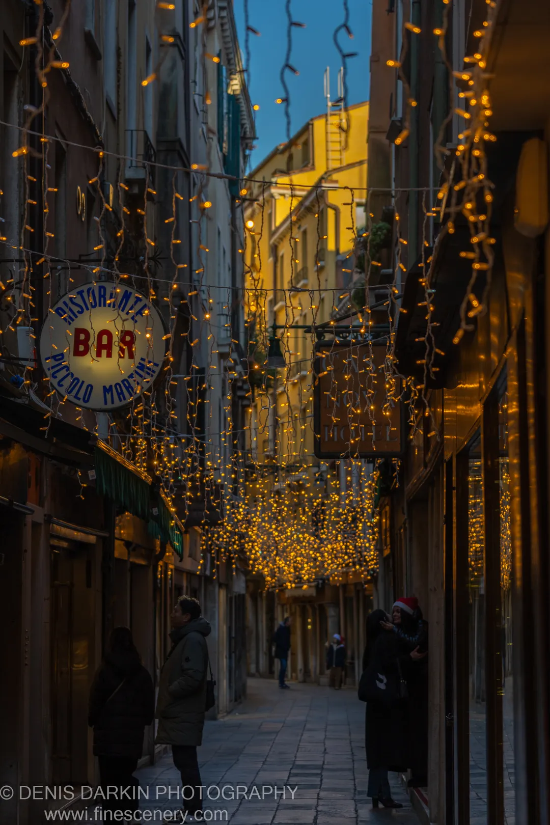 A narrow Venetian calle hung with warm fairy lights, figures in winter coats below a strip of blue morning sky.