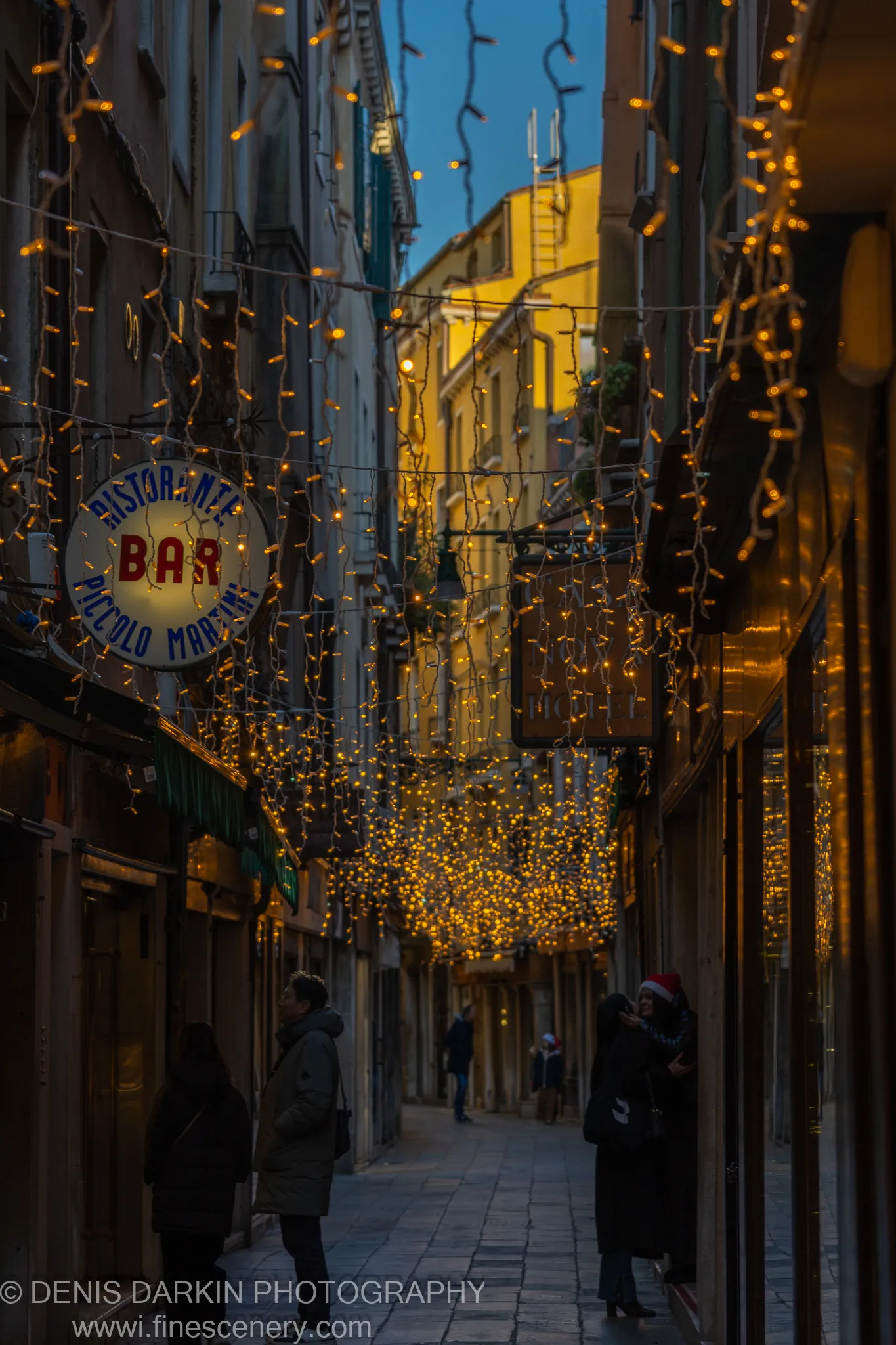 A narrow Venetian calle hung with warm fairy lights, figures in winter coats below a strip of blue morning sky.