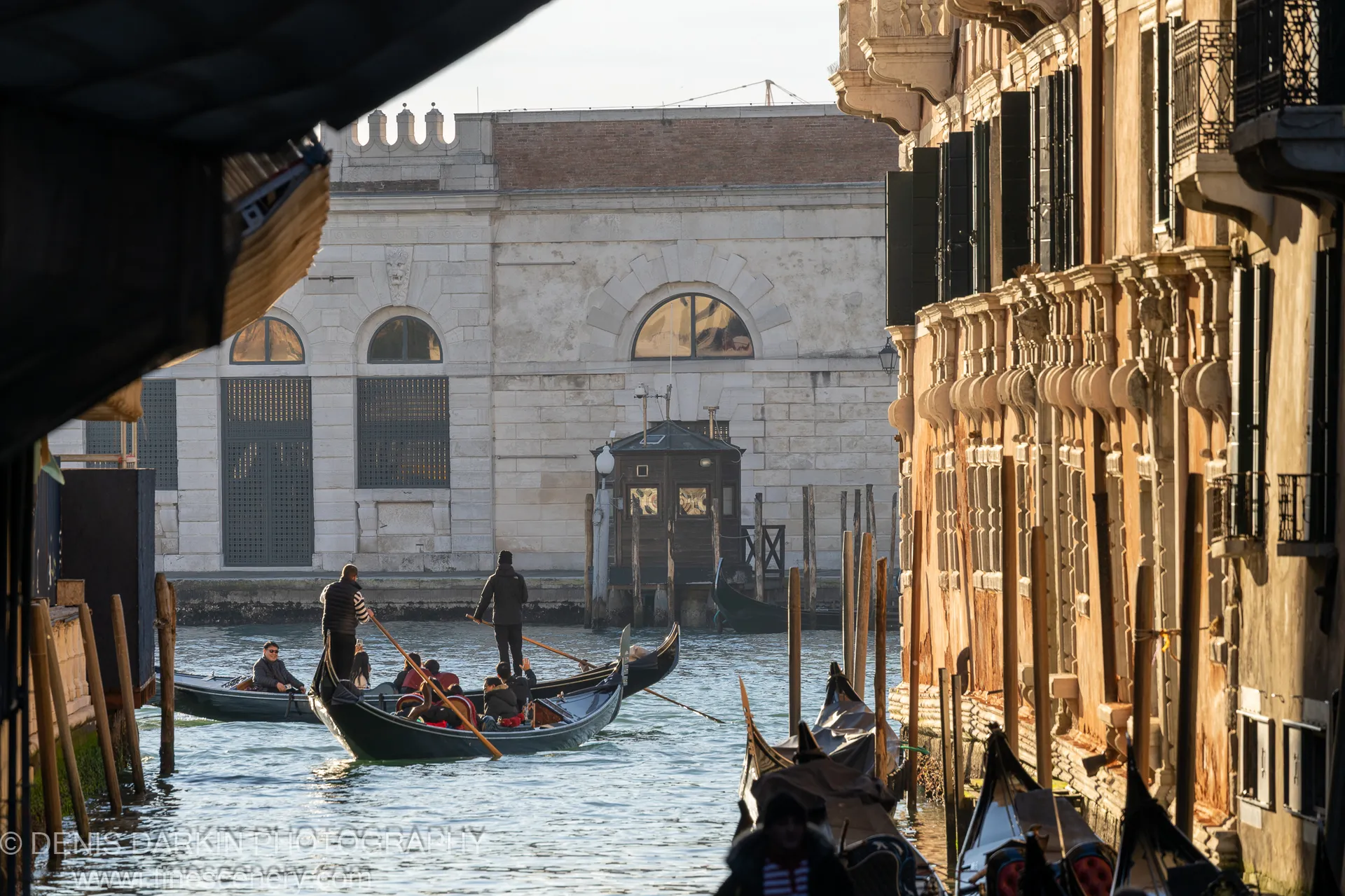Warm morning light falls on the stone facades of a Venice canal, gondolas moored between wooden poles in still water below.