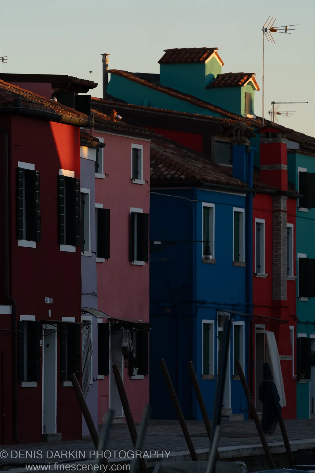 Blurred wooden pilings anchor the foreground as the painted facades of Burano rise in red, pink, and cobalt blue at dusk.