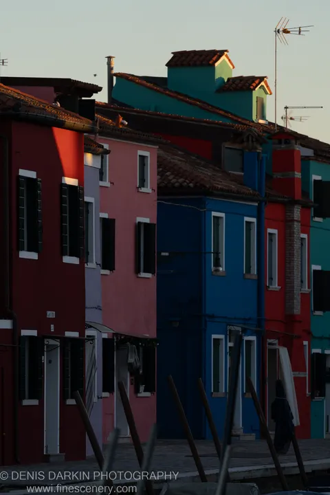 Blurred wooden pilings anchor the foreground as the painted facades of Burano rise in red, pink, and cobalt blue at dusk.