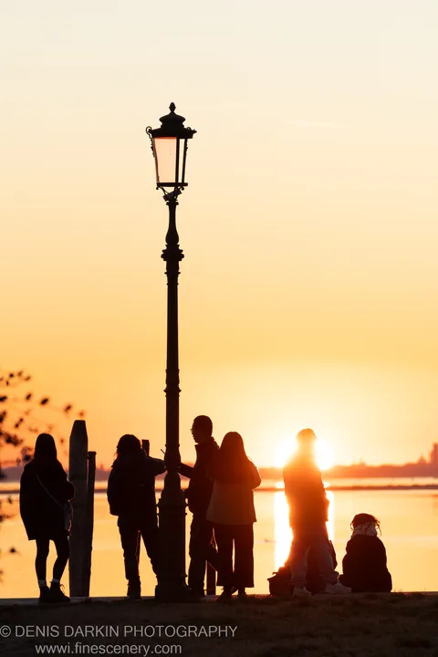 A vintage lamppost stands silhouetted against the low winter sun over the Burano waterfront, with warm light reflected in the water below.