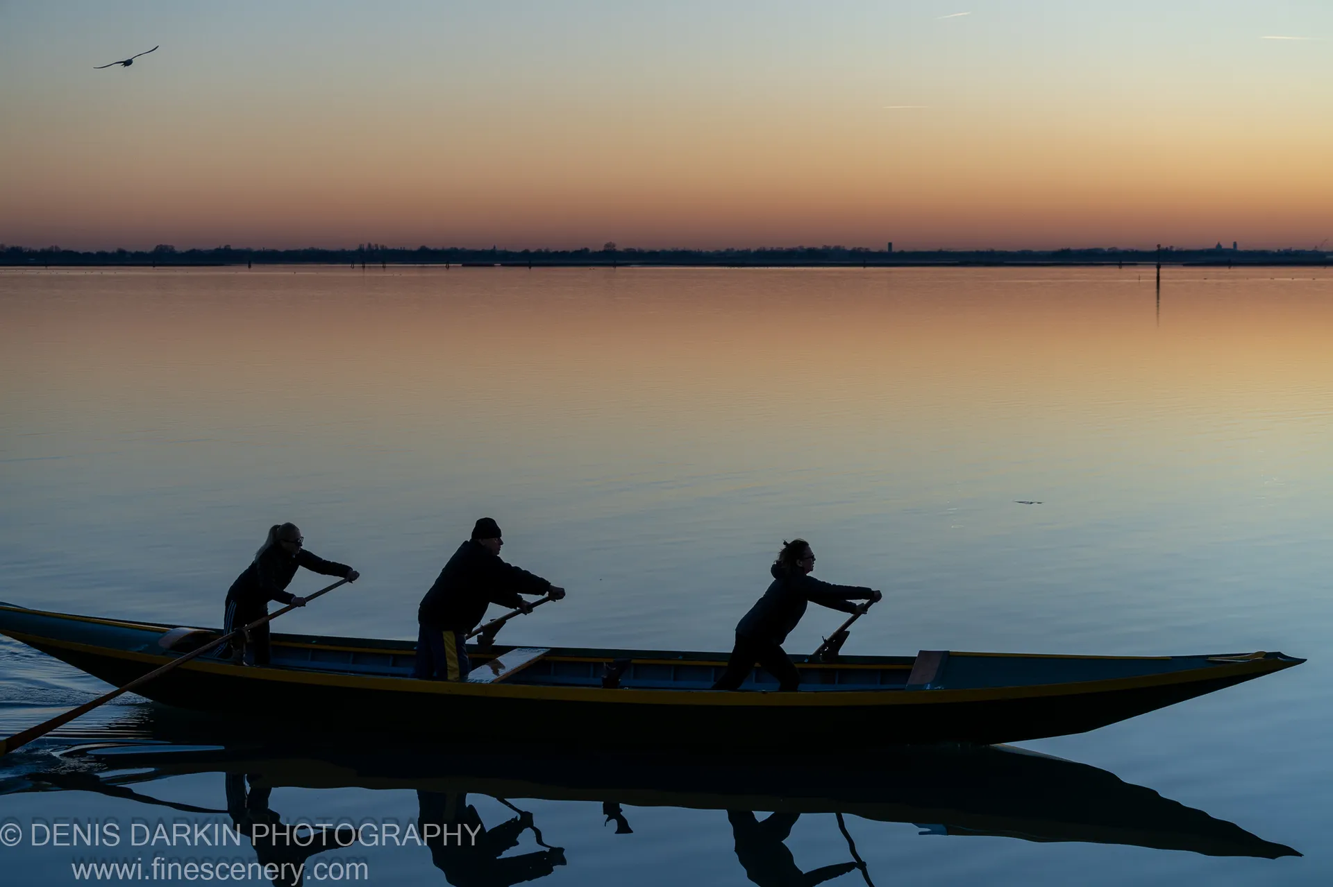 Silhouetted rowers power a long vessel across still water as the sky shifts from deep blue to peach at sunset near Burano.