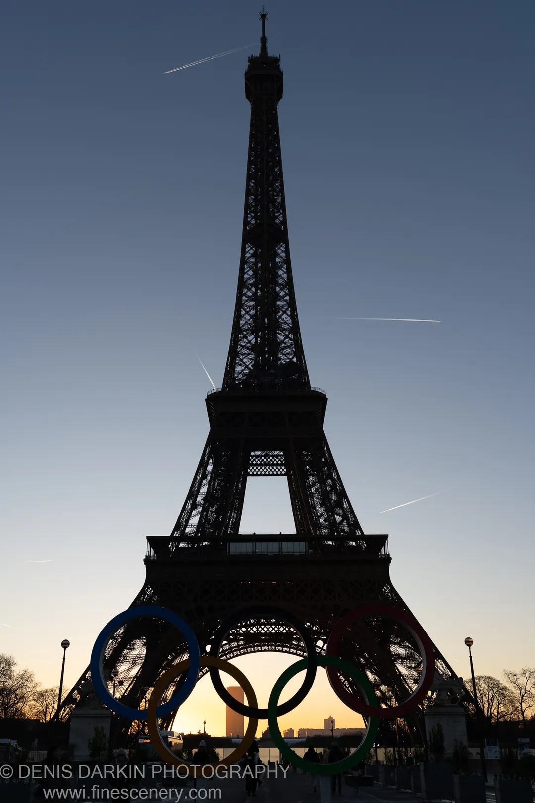 The Eiffel Tower rises as a dark lattice silhouette against a deep blue winter sky at dawn, with Olympic rings in the foreground.