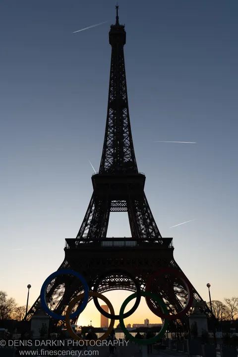 The Eiffel Tower rises as a dark lattice silhouette against a deep blue winter sky at dawn, with Olympic rings in the foreground.