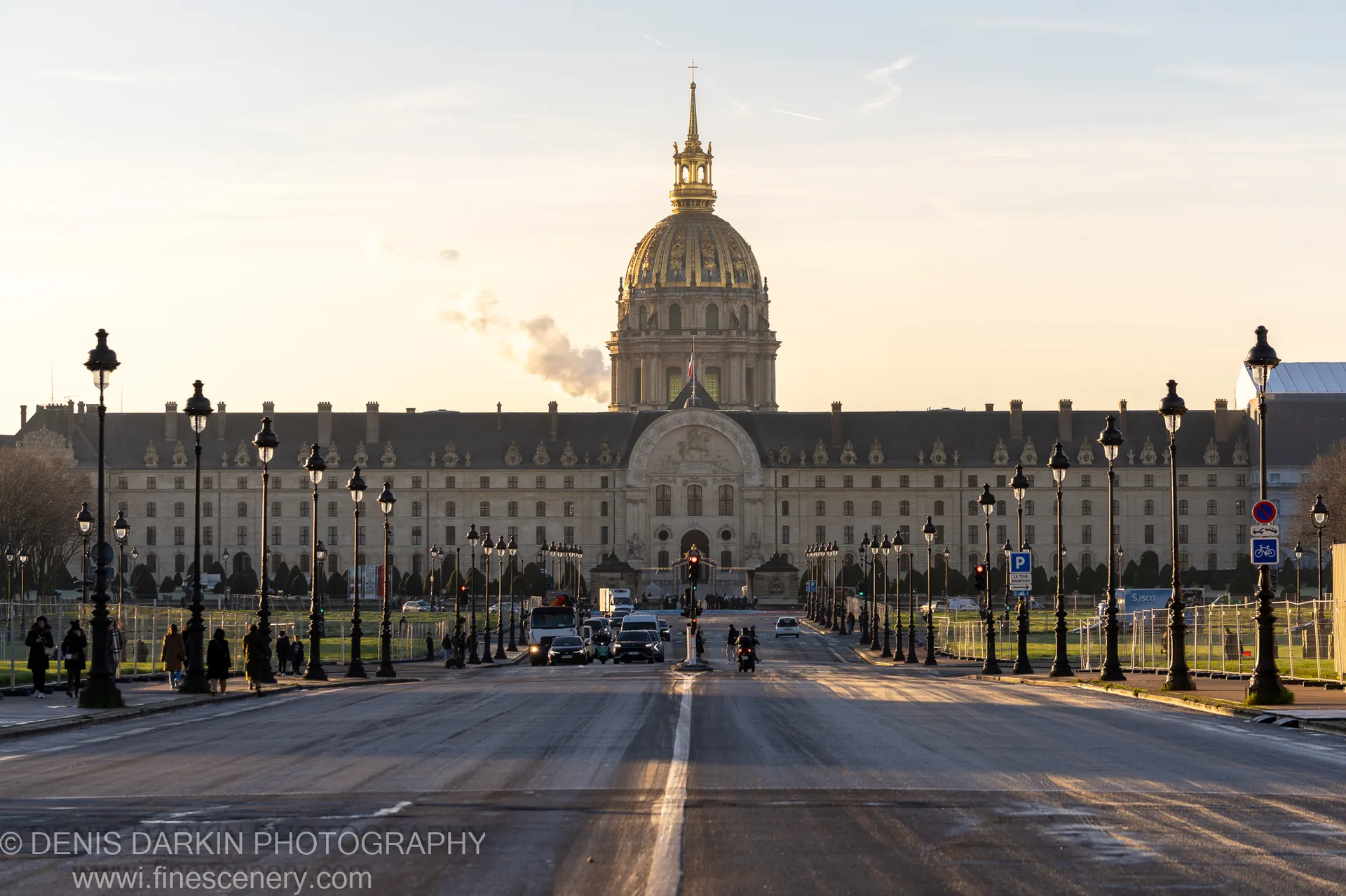 Black lampposts recede along a wide winter avenue in Paris, converging toward a golden dome silhouetted against a pale hazy sky.