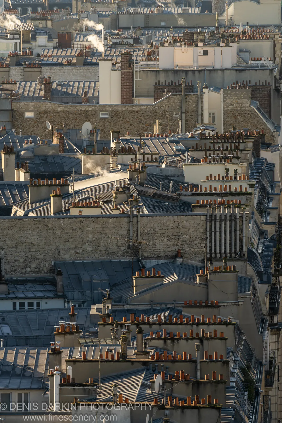 Wisps of steam rise from chimney flues above the dense Parisian slate rooftops, stone facades and satellite dishes visible below.