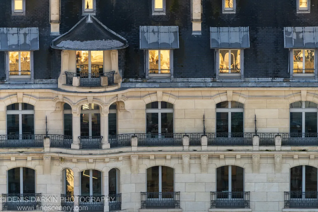 Warm winter light from upper dormer windows contrasts with shadowed slate below, wrought iron railings and arched stone openings across the Paris facade.