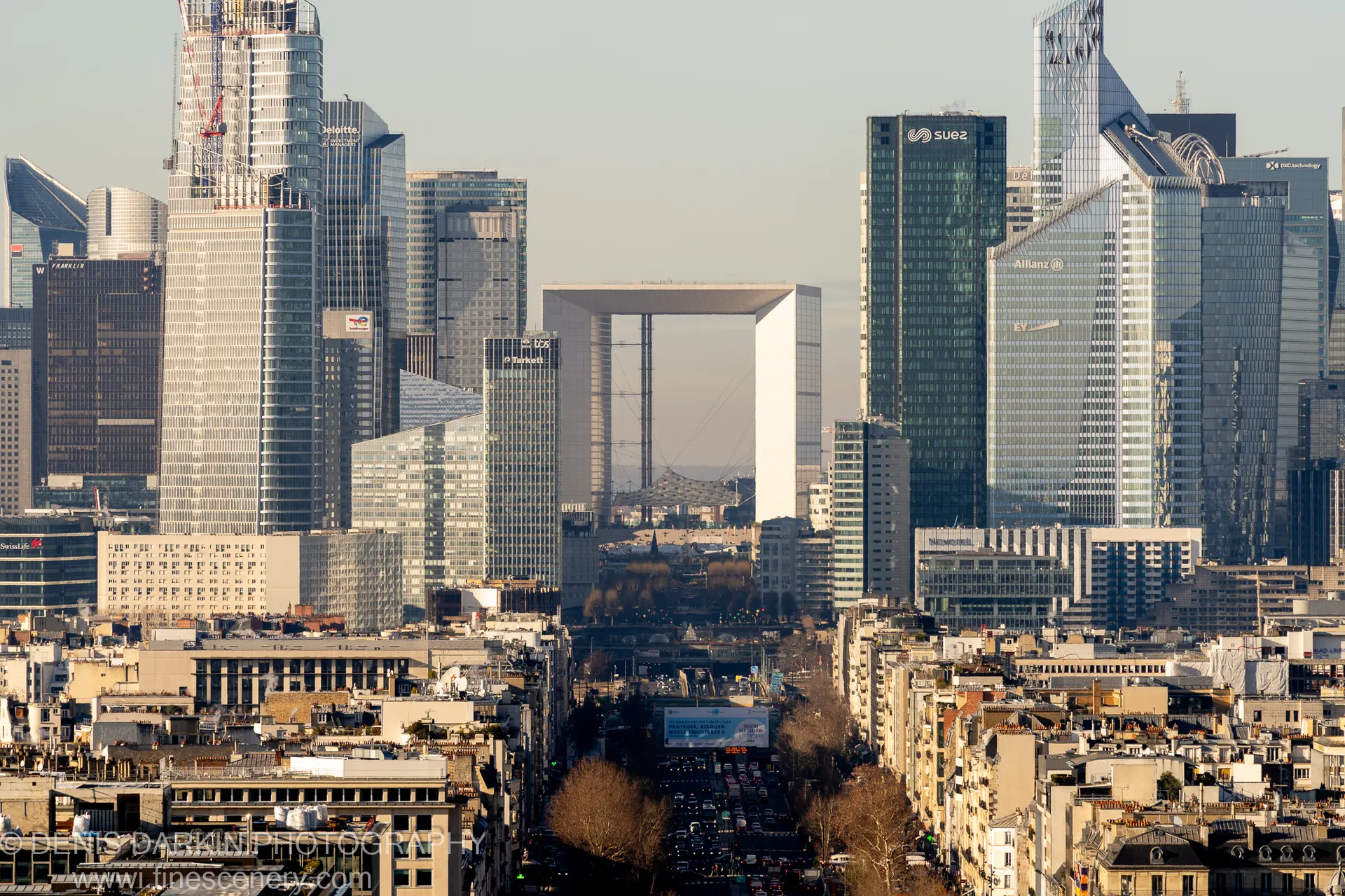 Telephoto compression merges Parisian slate rooftops with distant glass towers, a white arch structure standing at the center of the cluster in winter light.