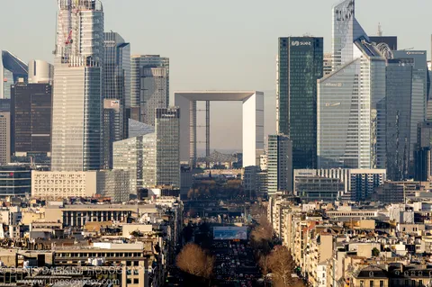 Telephoto compression merges Parisian slate rooftops with distant glass towers, a white arch structure standing at the center of the cluster in winter light.