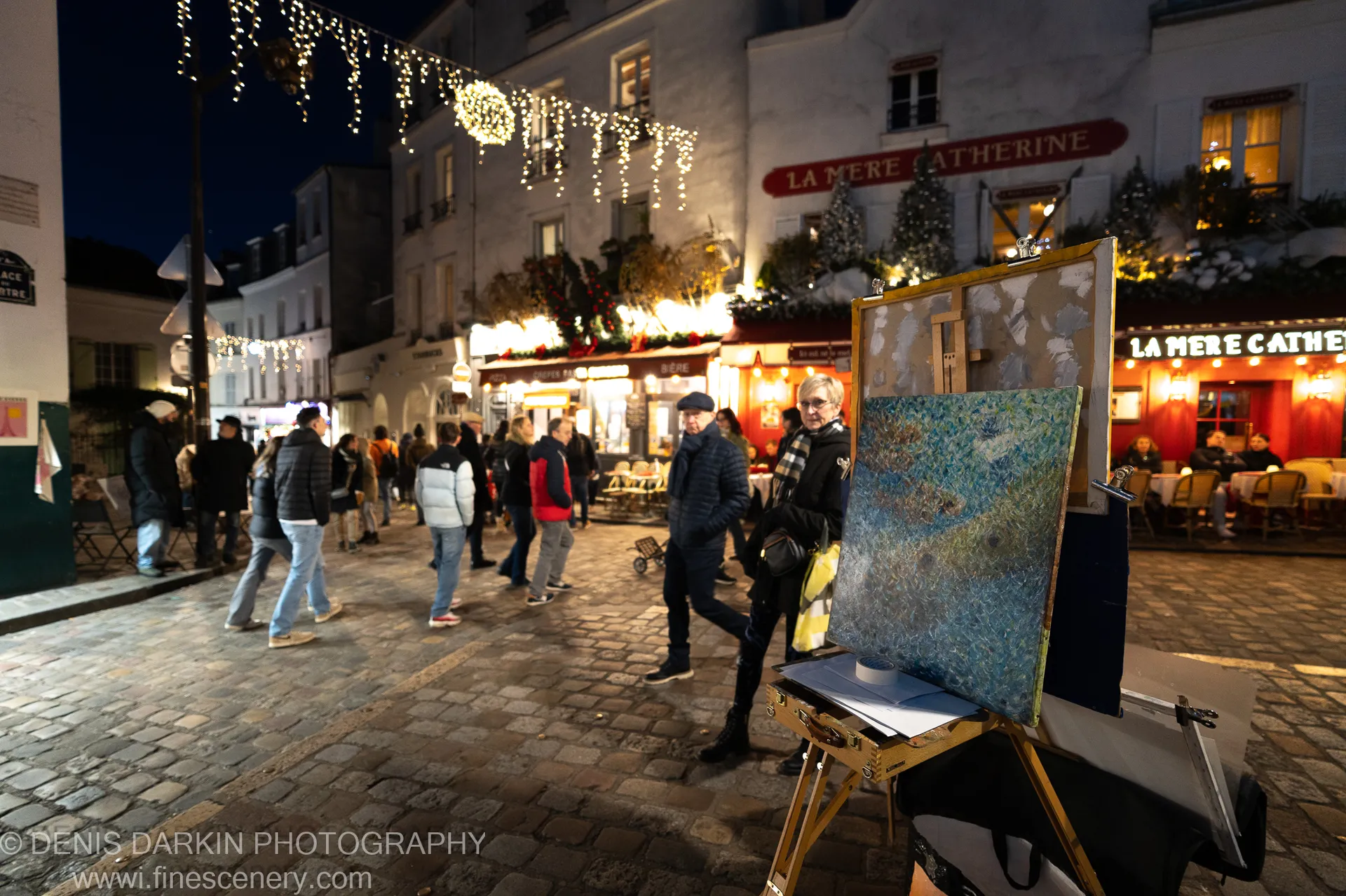 A painter's easel stands in a cobblestone Paris square at dusk, warm fairy lights strung overhead and winter pedestrians passing by cafe storefronts.