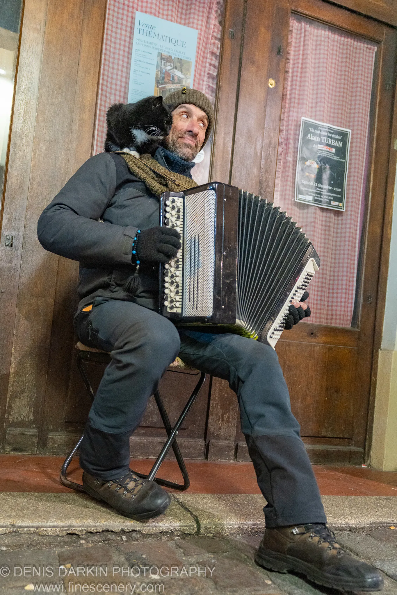 A street musician plays accordion outside a Paris doorway with a black cat resting on his shoulder, red checkered curtains behind him.