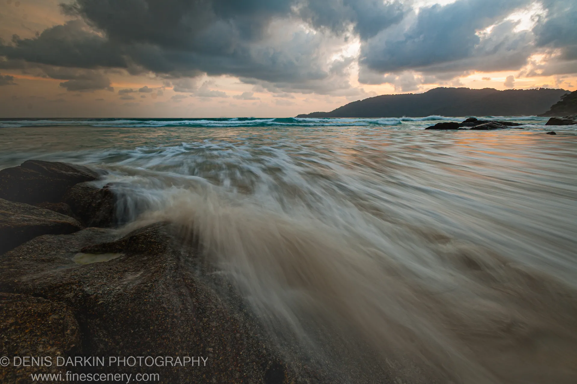 Warm orange light filters through heavy clouds near the horizon, casting a glow across a long-exposure seascape and wet rocks on the Malaysian coast.