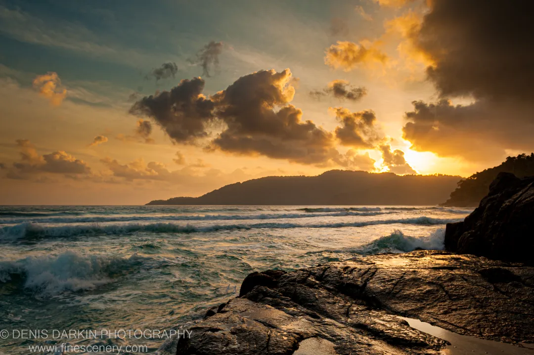 Sunburst light breaks through clouds at sunrise, warm reflections on wet rocks in the foreground, white-capped waves and a distant headland, Malaysian coast.