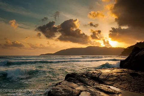 Sunburst light breaks through clouds at sunrise, warm reflections on wet rocks in the foreground, white-capped waves and a distant headland, Malaysian coast.