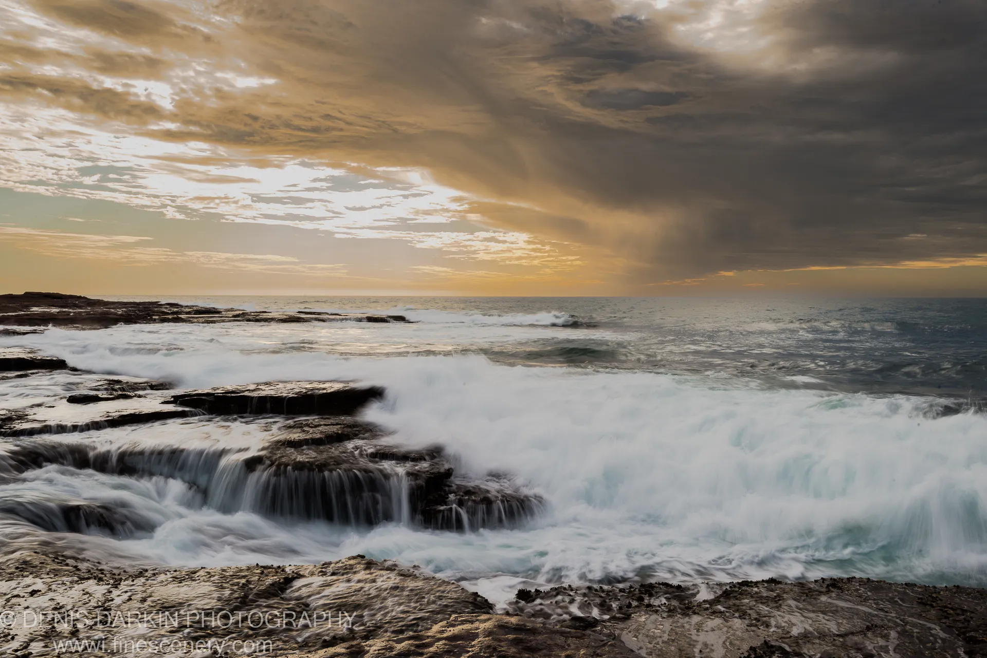 Dark wet rocks with white foam in the foreground and warm light on the edge of heavy storm clouds above the Western Australian coast.
