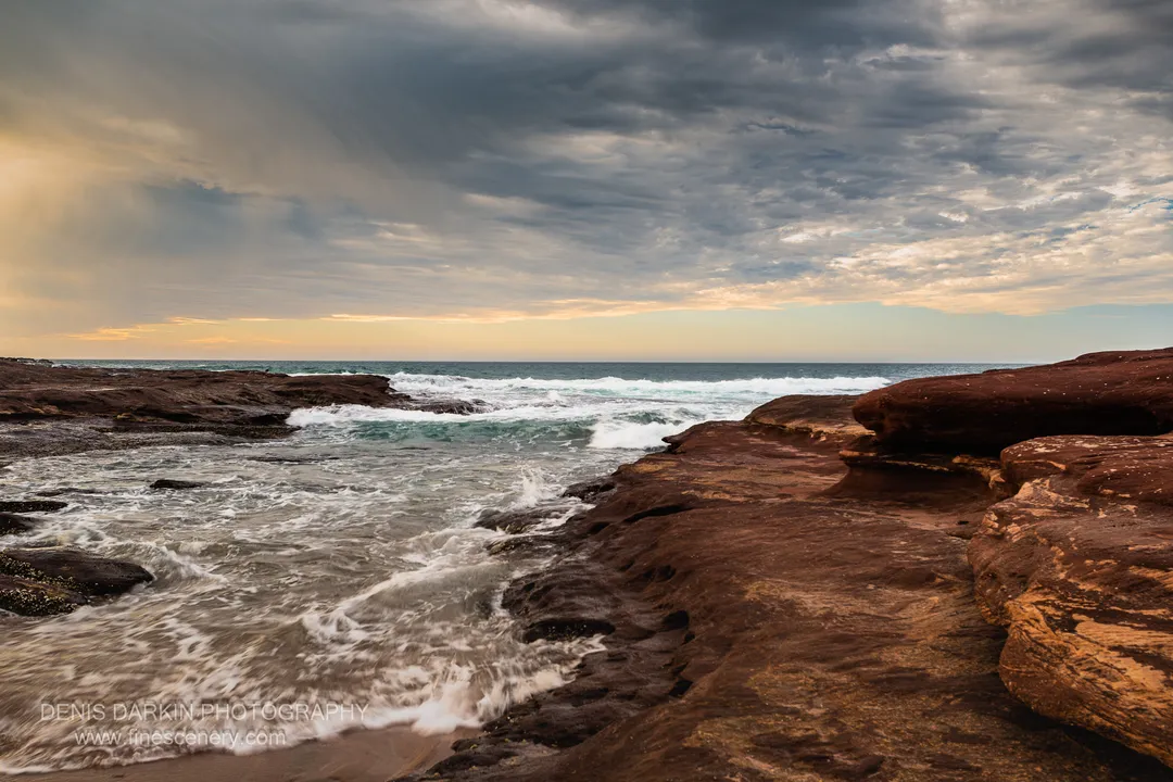 Warm light breaks through grey clouds above layered red sandstone ledges on the Western Australian coast, with turbulent white surf in the foreground.