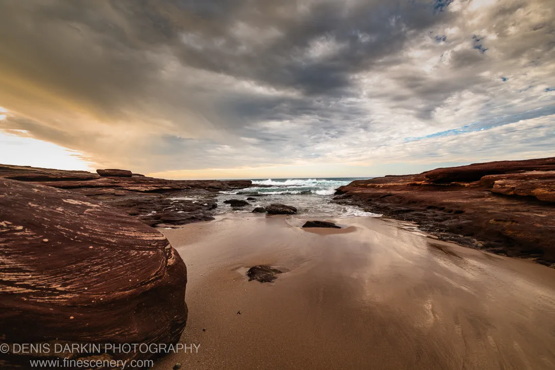Flat sandstone platform on the Western Australian coast, its wet surface reflecting dark storm clouds and a narrow band of warm light at the horizon.