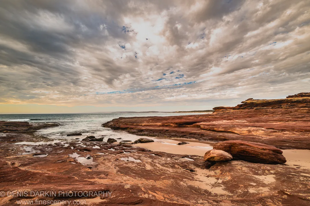 Reddish-brown sandstone curving around a small sandy inlet on the Western Australian coast, with tidal pools and heavy clouds above.