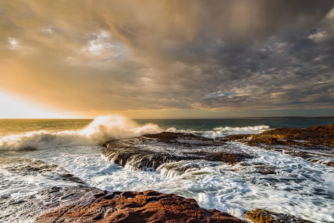 Turbulent white water over dark rock ledges on the Western Australian coast, warm light breaking through storm clouds overhead.