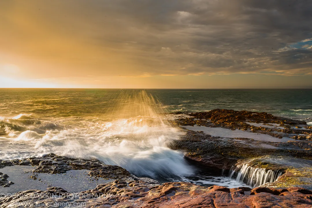 Backlit sea spray rising over moss-covered red rock shelves on the Western Australian coast, with storm clouds above at dusk.