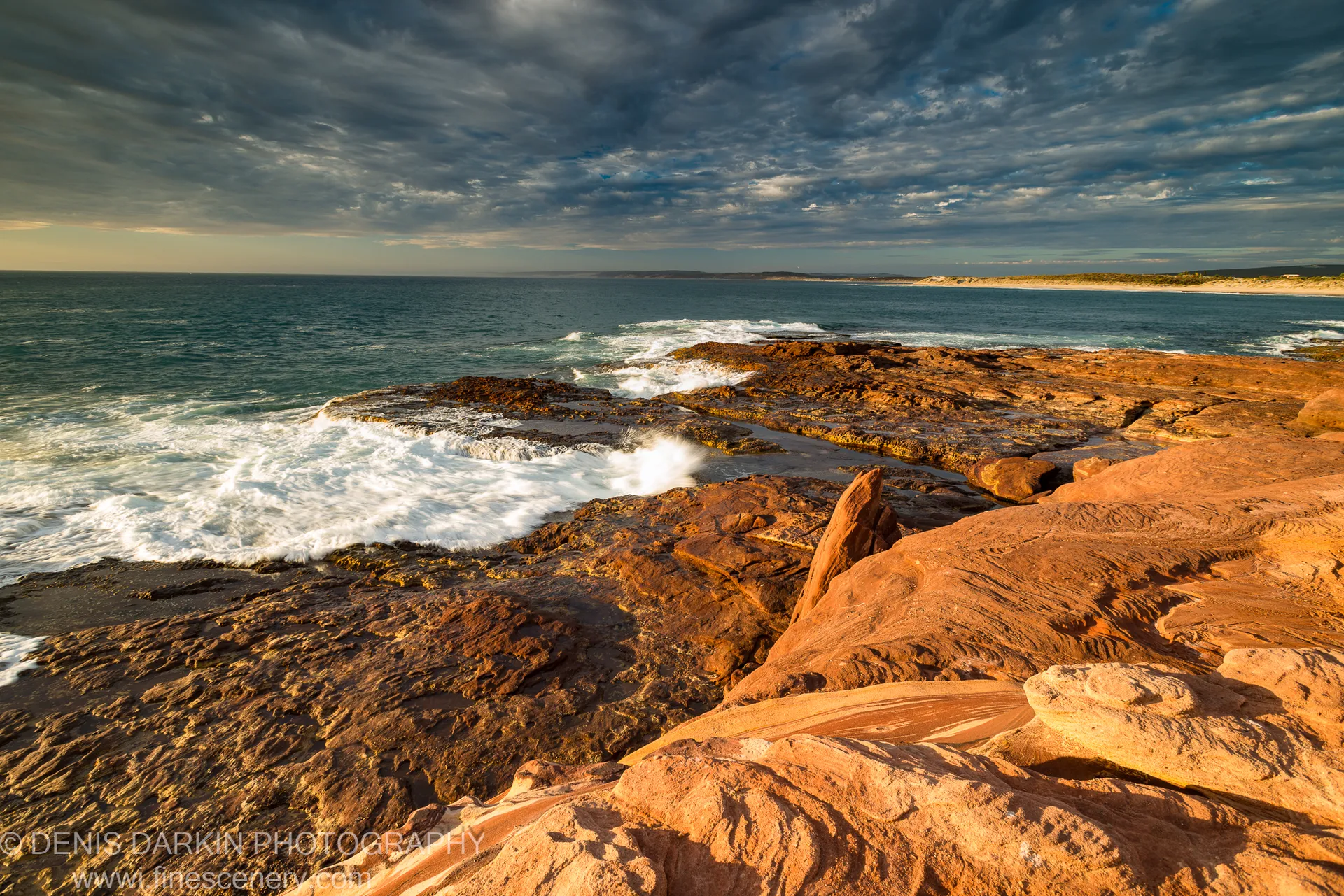 Deep red eroded rock formations on the Western Australian coast, white surf breaking at their base beneath dark storm clouds at dusk.