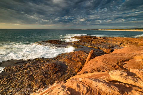Deep red eroded rock formations on the Western Australian coast, white surf breaking at their base beneath dark storm clouds at dusk.
